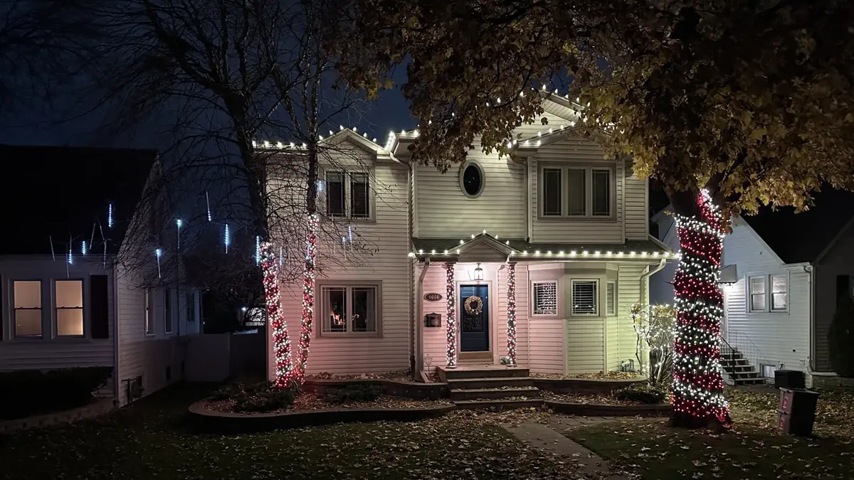 Warm white C9 roofline with lit red and white garland wrapped around the porch columns and trees, and a lit wreath at the front door