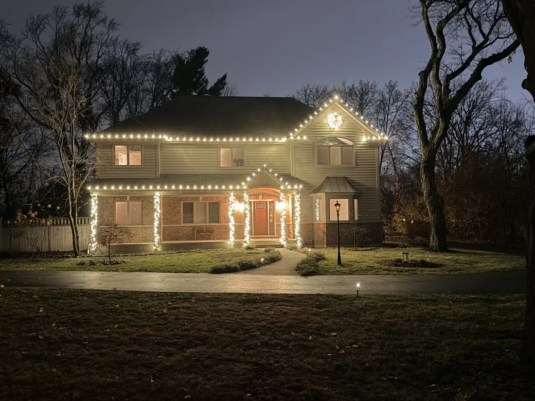 Two-story colonial in Prospect Heights at night with warm white C9 lights along the roofline and gables, lit garland on the porch columns, and a lit wreath on the front gable