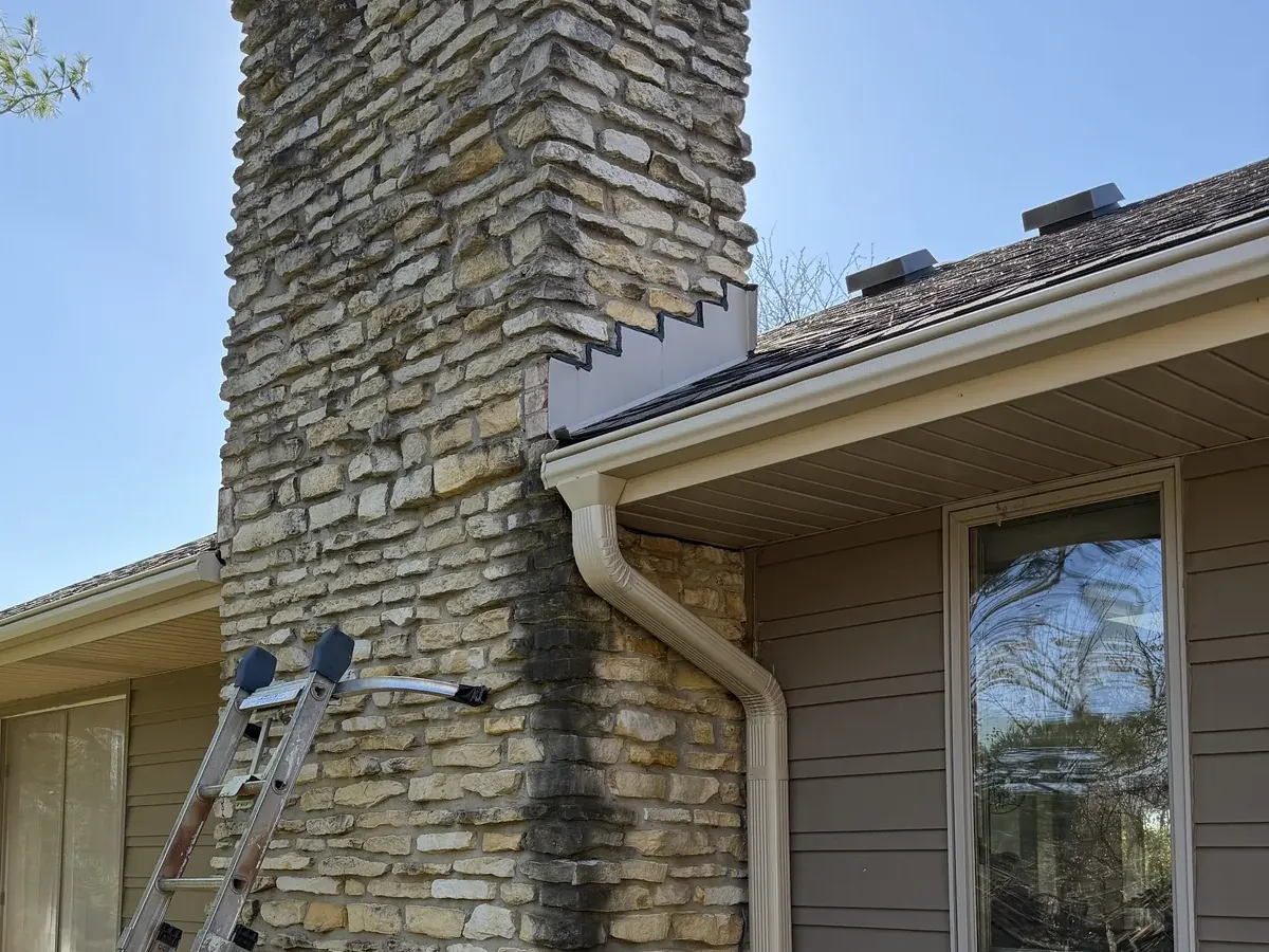A stone chimney and taupe lap siding before washing, dark algae streaking down the chimney corner